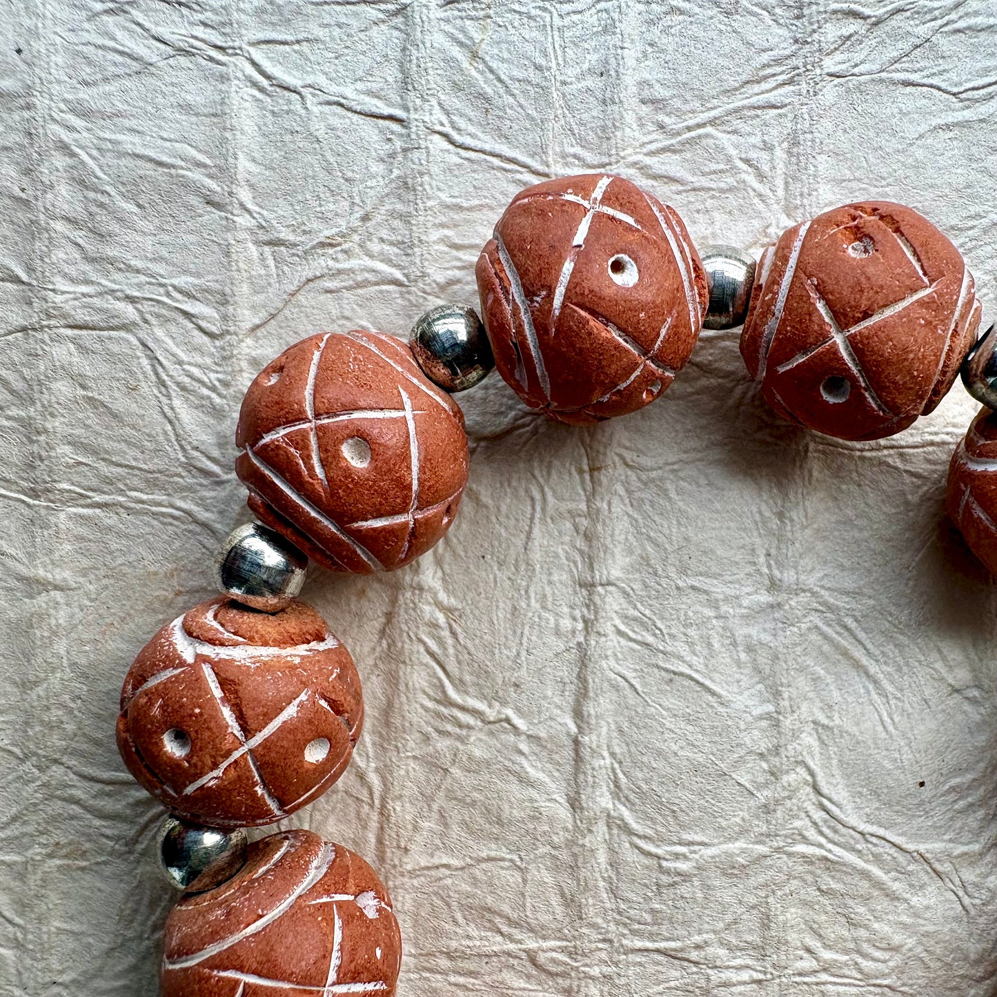 A close-up of terracotta-colored ceramic beads with white painted geometric lines and small holes, separated by shiny silver metal beads, laid on a textured off-white surface.