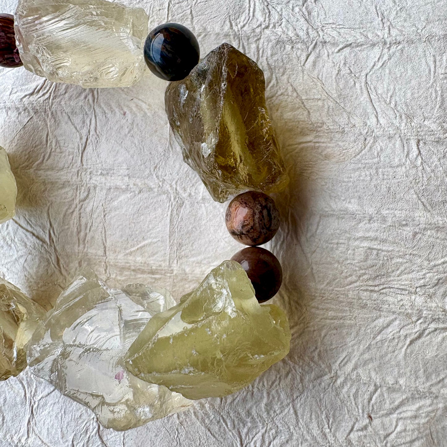 A handmade beaded bracelet featuring raw, translucent yellow-green quartz crystal chunks and purple beads, arranged in a circle on a textured off-white paper background.