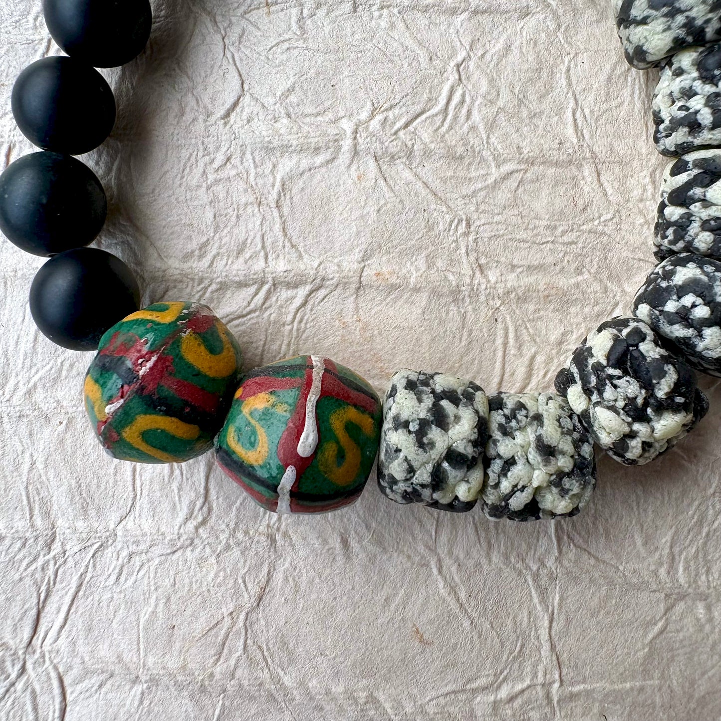 The bracelet features matte black round beads, polished black stone beads, a cluster of black-and-white marbled beads, and two colorful patterned beads with green, red, and yellow details.