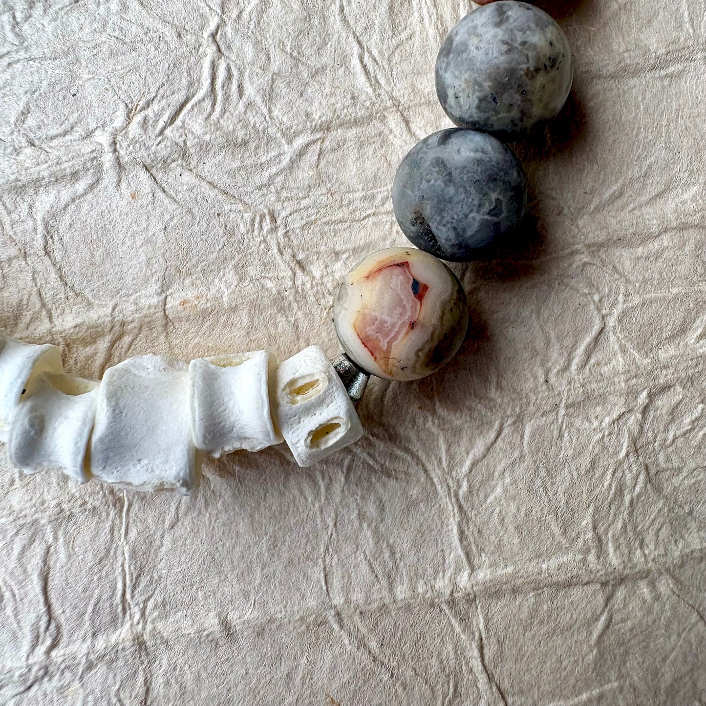 A close-up of a beaded strand featuring white bone-like cylindrical beads, dark gray stone round beads, and one unique marbled bead with pink and cream patterns.