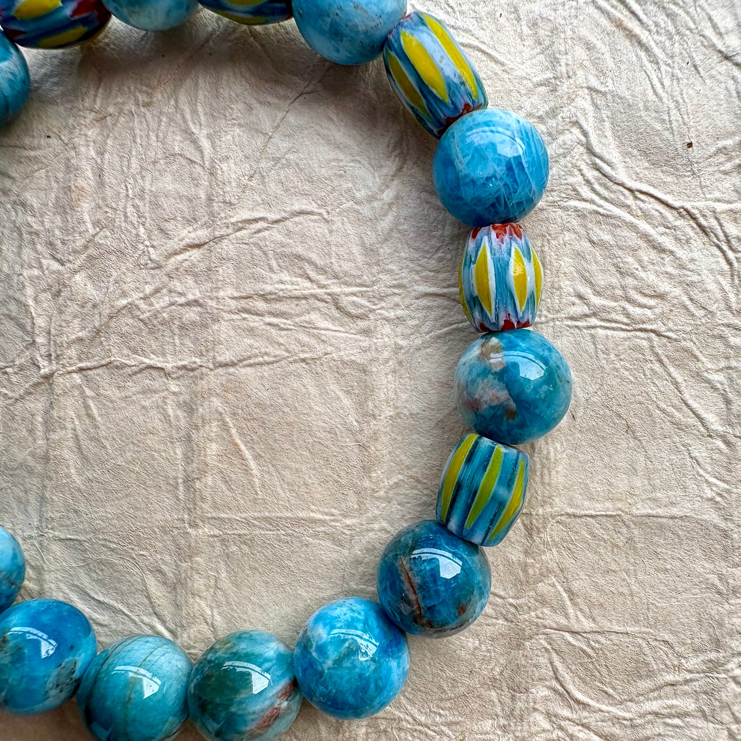 A close-up of a beaded bracelet made of glossy turquoise-blue beads, including round beads with marbled blue and white patterns and striped beads with yellow, blue, and red accents.