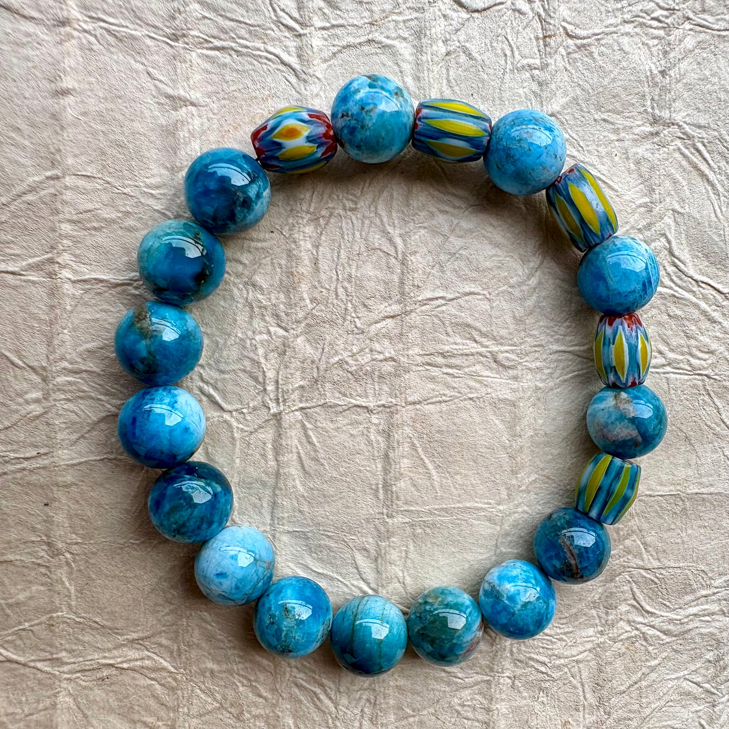 A beaded stretch bracelet made of glossy blue marbled beads and a few striped accent beads in yellow, green, and red, displayed on a textured beige fabric background.