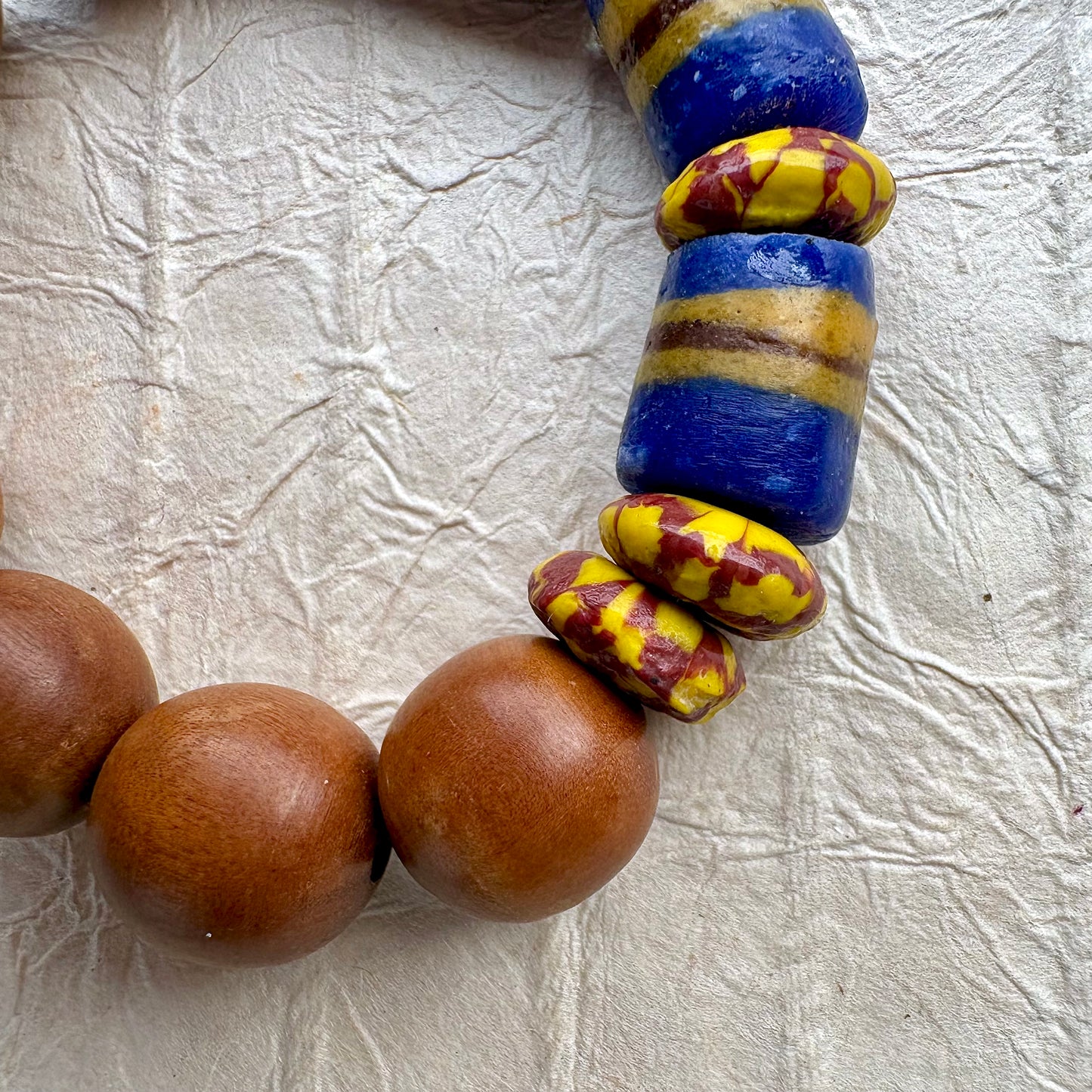 A close-up of a beaded necklace or bracelet featuring smooth round brown wooden beads and glossy blue glass beads with yellow and reddish-brown patterned accents, laid on a textured off-white surface.