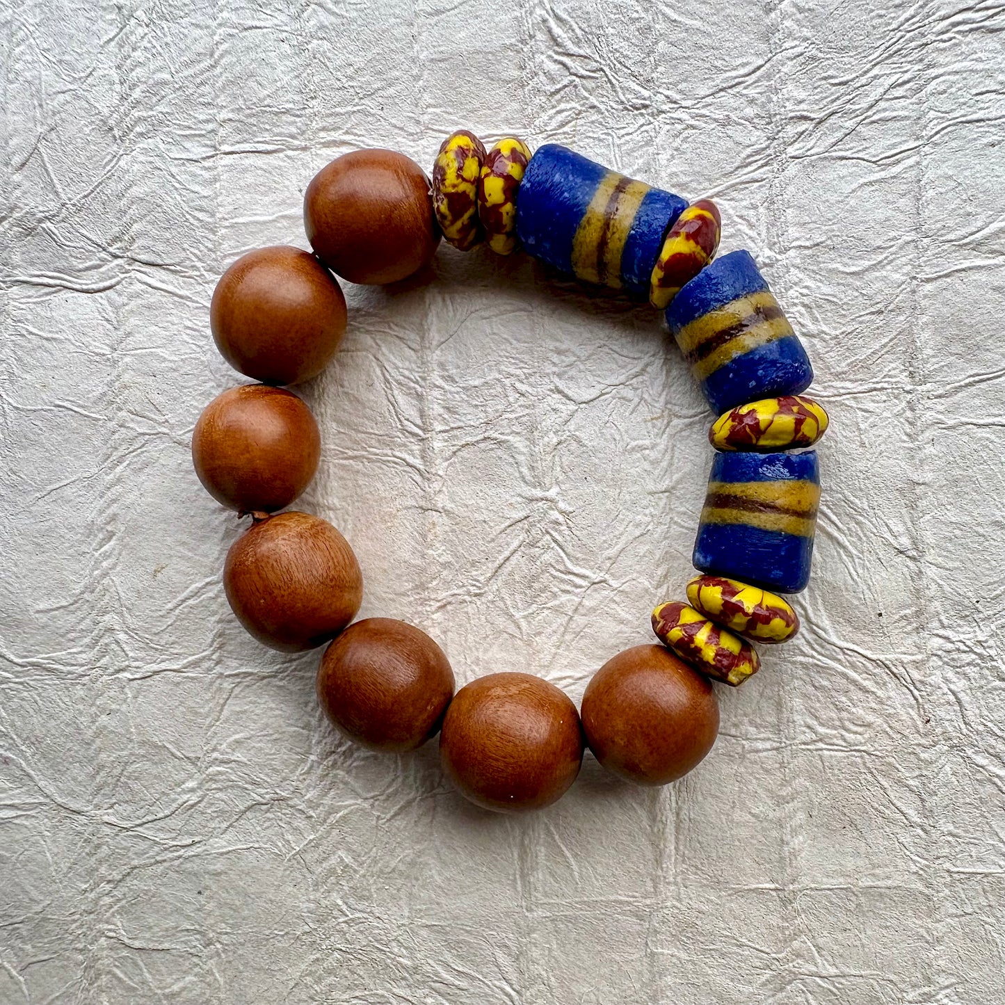 A beaded bracelet made of round medium-brown wooden beads and two blue glass cylinder beads with yellow stripes, accented by small yellow and red patterned beads, displayed on a textured light gray background.