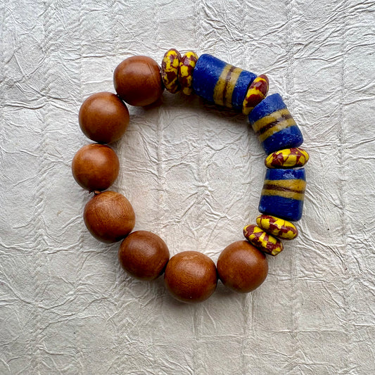 A beaded bracelet made of round medium-brown wooden beads and two blue glass cylinder beads with yellow stripes, accented by small yellow and red patterned beads, displayed on a textured light gray background.