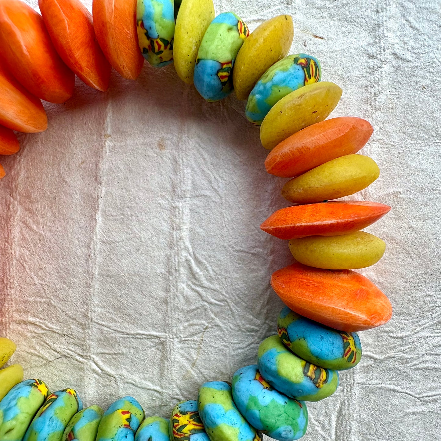 A handmade beaded bracelet made of flat, elongated beads in orange, yellow, and turquoise blue with brown marbling, arranged in a circle on a textured white background.