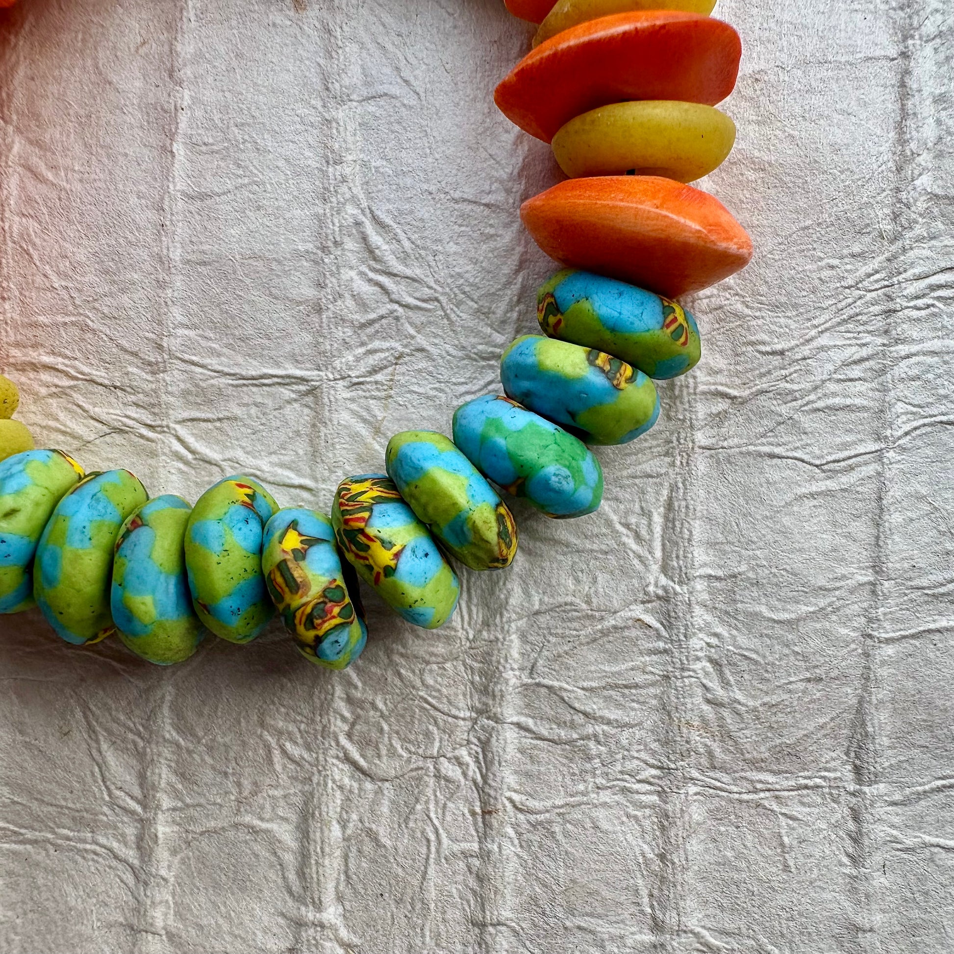 A handmade beaded bracelet made of flat, elongated beads in orange, yellow, and turquoise blue with brown marbling, arranged in a circle on a textured white background.