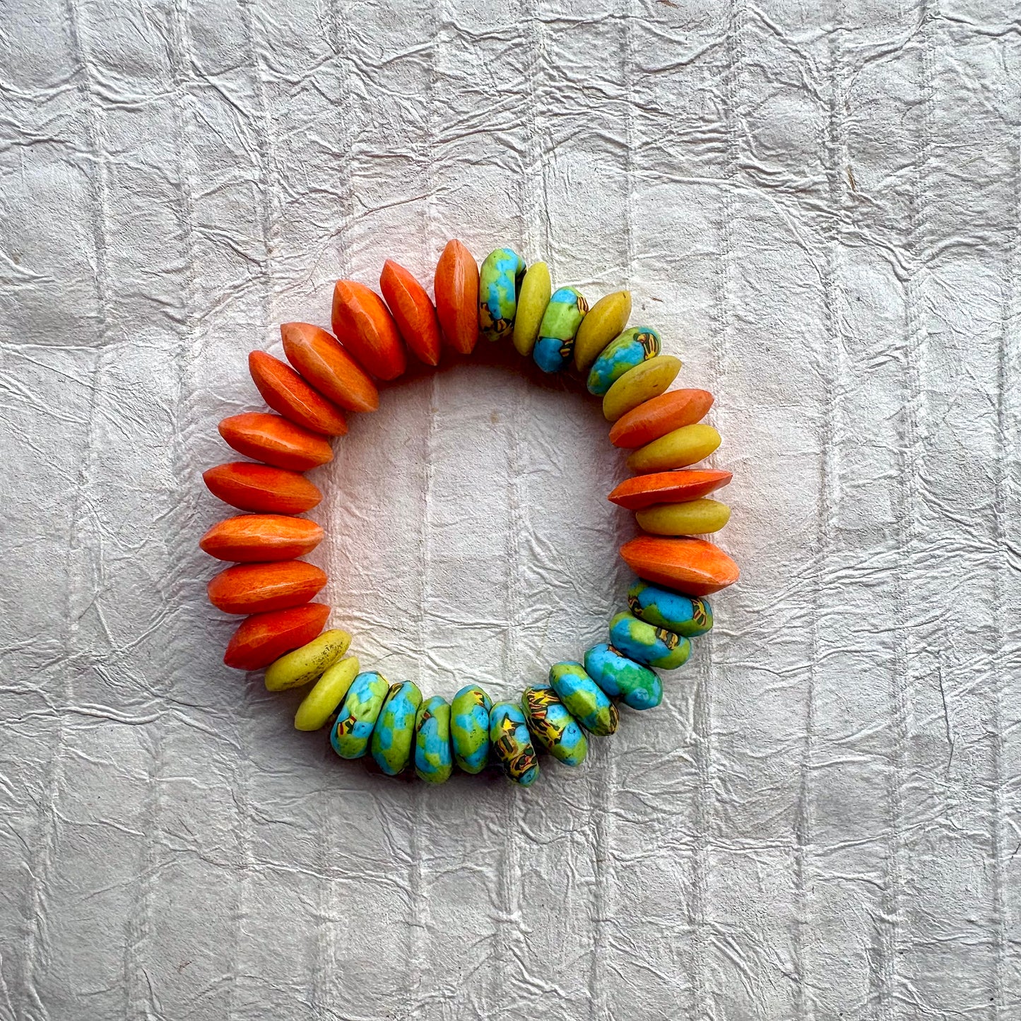 A handmade beaded bracelet made of flat, elongated beads in orange, yellow, and turquoise blue with brown marbling, arranged in a circle on a textured white background.