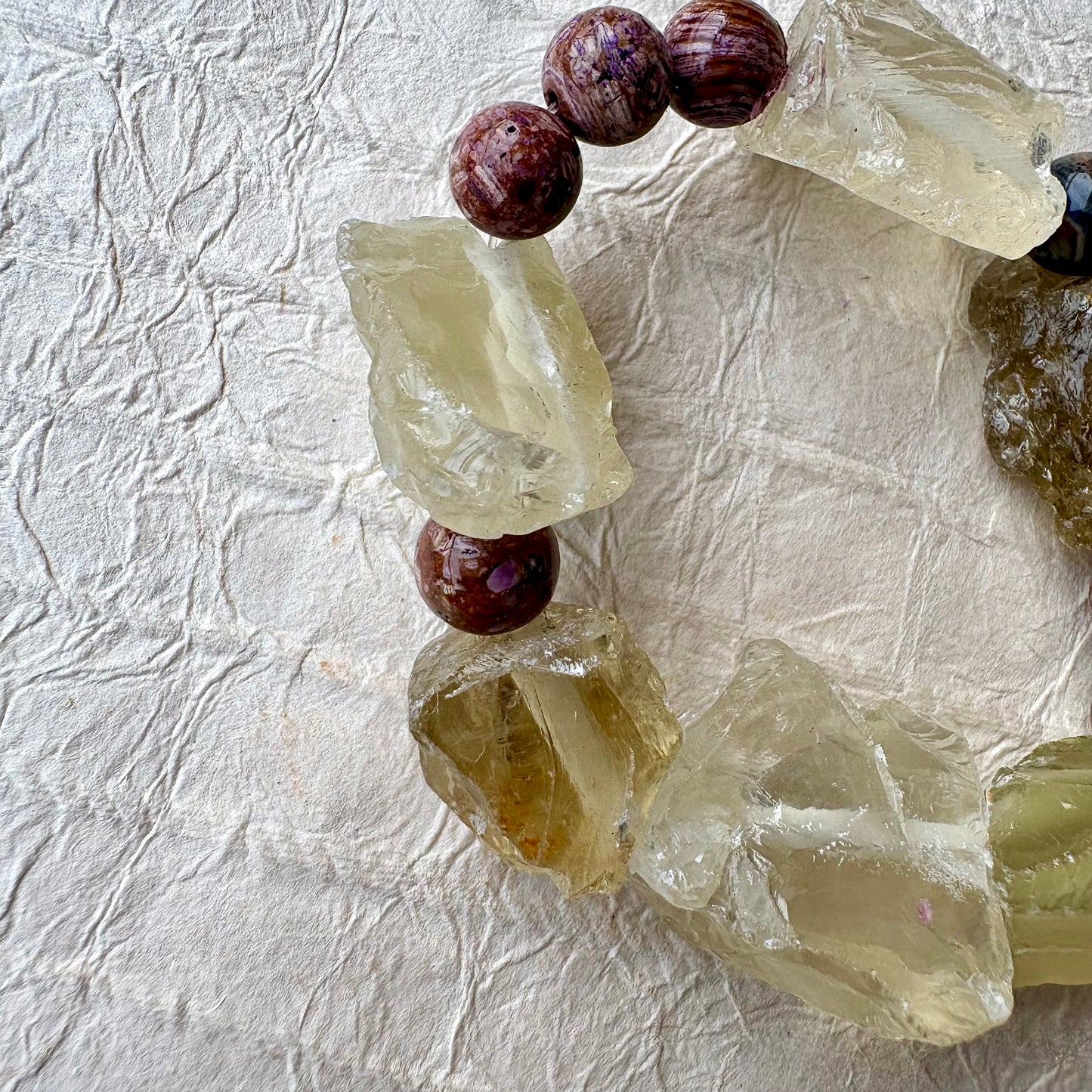 A handmade beaded bracelet featuring raw, translucent yellow-green quartz crystal chunks and purple beads, arranged in a circle on a textured off-white paper background.