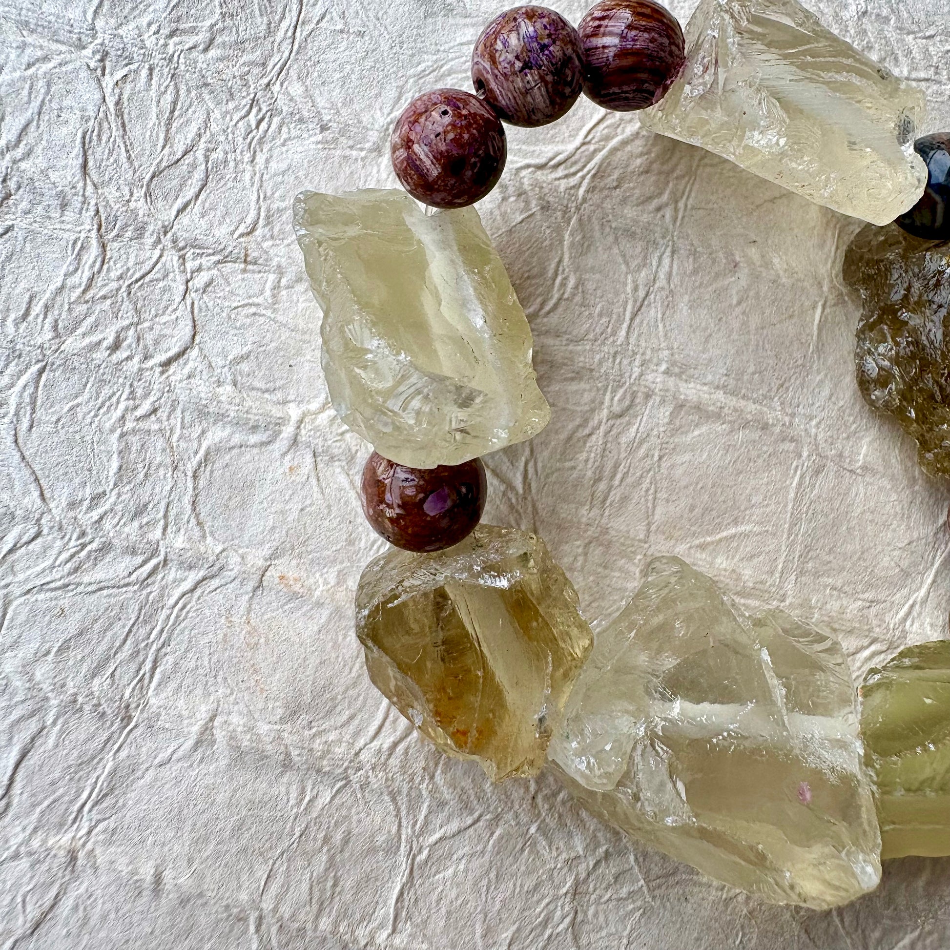 A handmade beaded bracelet featuring raw, translucent yellow-green quartz crystal chunks and purple beads, arranged in a circle on a textured off-white paper background.