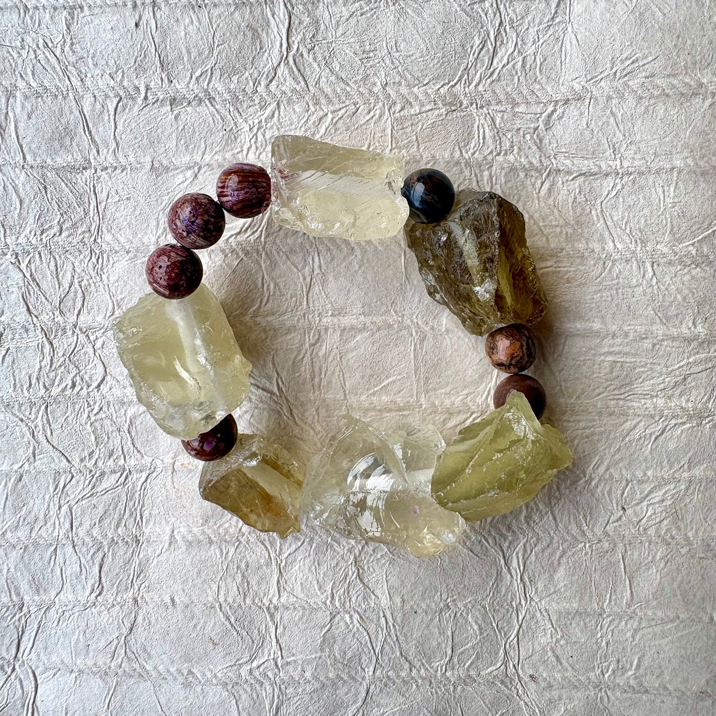 A handmade beaded bracelet featuring raw, translucent yellow-green quartz crystal chunks and purple beads, arranged in a circle on a textured off-white paper background.