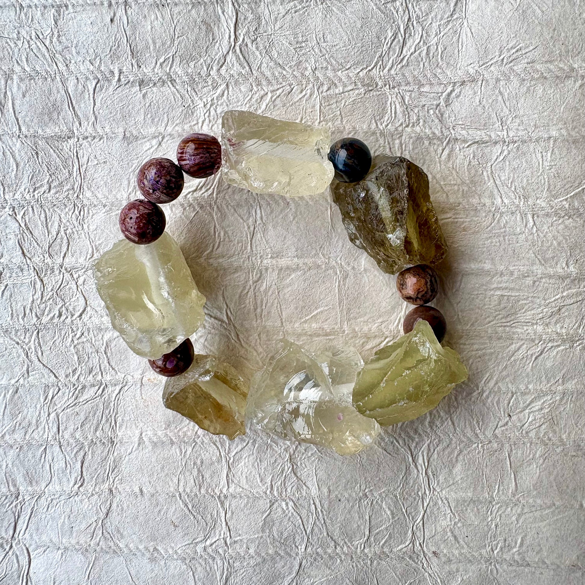 A handmade beaded bracelet featuring raw, translucent yellow-green quartz crystal chunks and purple beads, arranged in a circle on a textured off-white paper background.