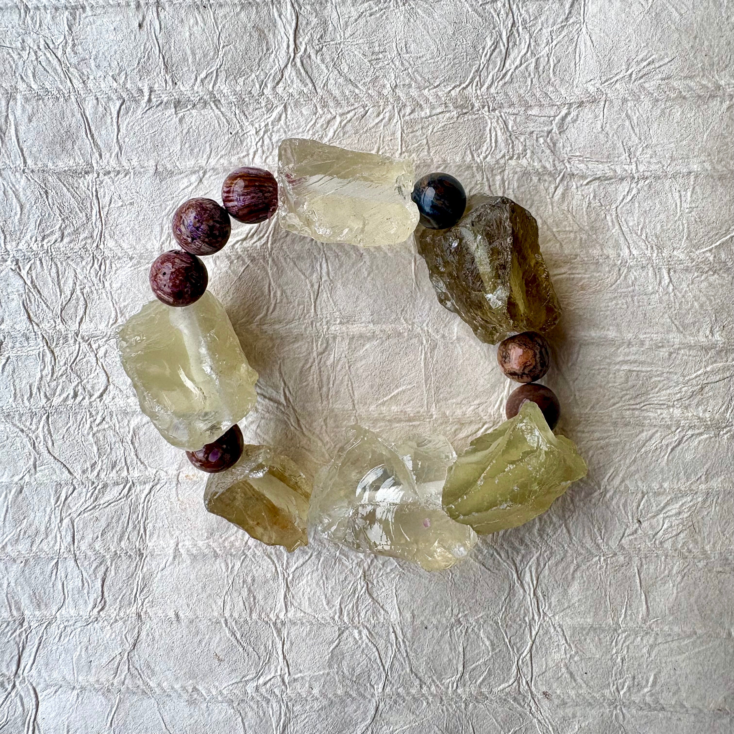 A handmade beaded bracelet featuring raw, translucent yellow-green quartz crystal chunks and purple beads, arranged in a circle on a textured off-white paper background.