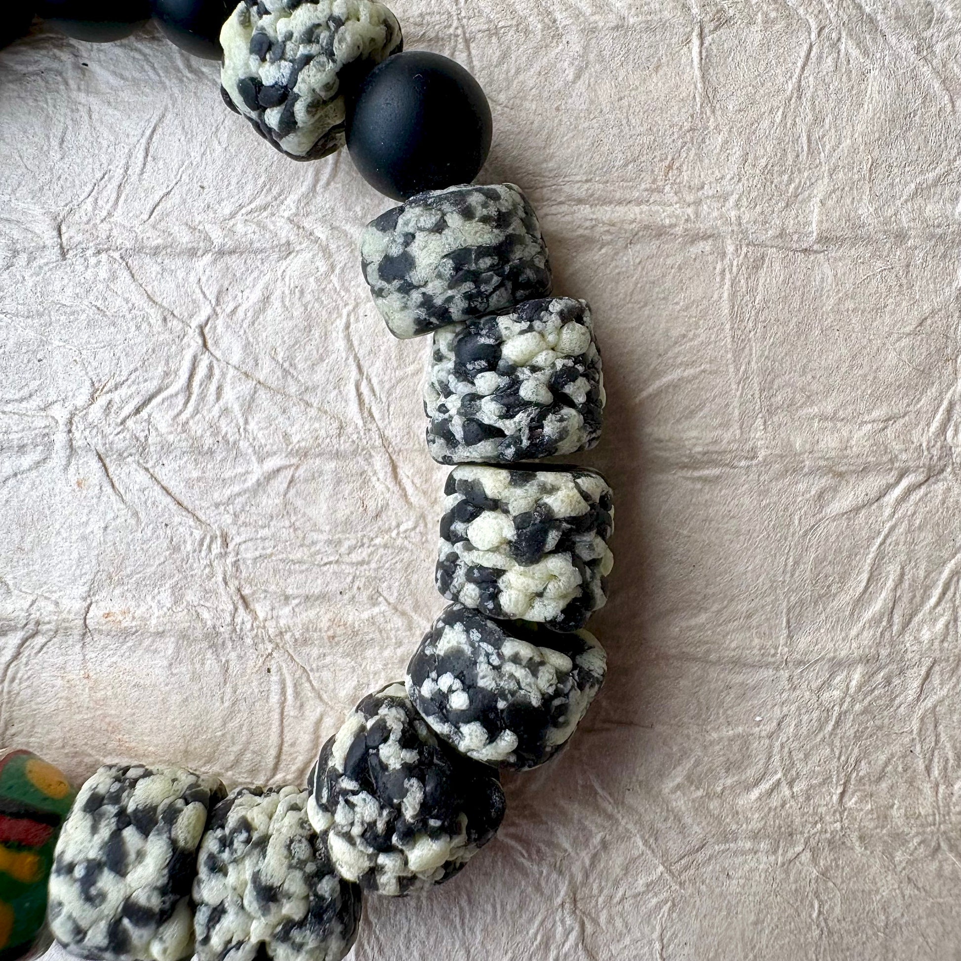 The bracelet features matte black round beads, polished black stone beads, a cluster of black-and-white marbled beads, and two colorful patterned beads with green, red, and yellow details.