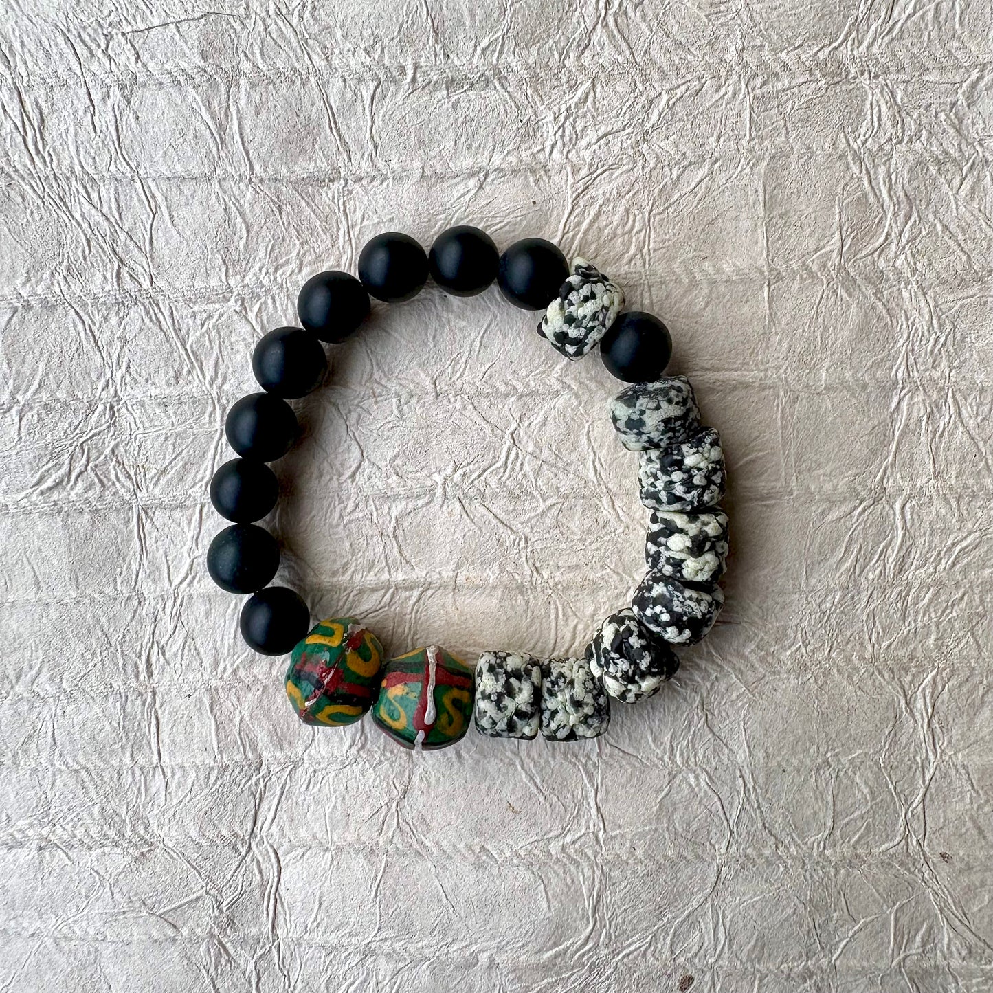 The bracelet features matte black round beads, polished black stone beads, a cluster of black-and-white marbled beads, and two colorful patterned beads with green, red, and yellow details.
