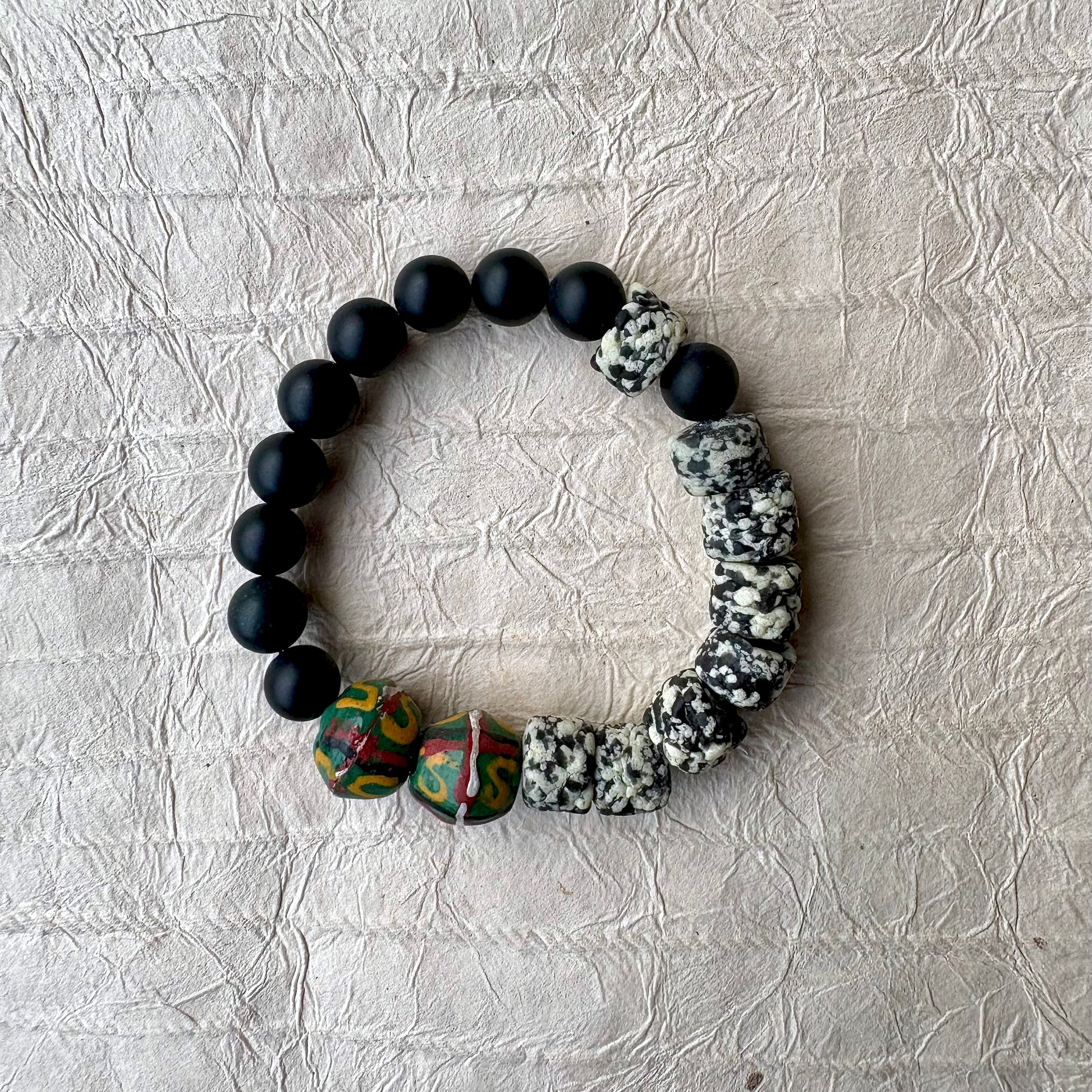 The bracelet features matte black round beads, polished black stone beads, a cluster of black-and-white marbled beads, and two colorful patterned beads with green, red, and yellow details.