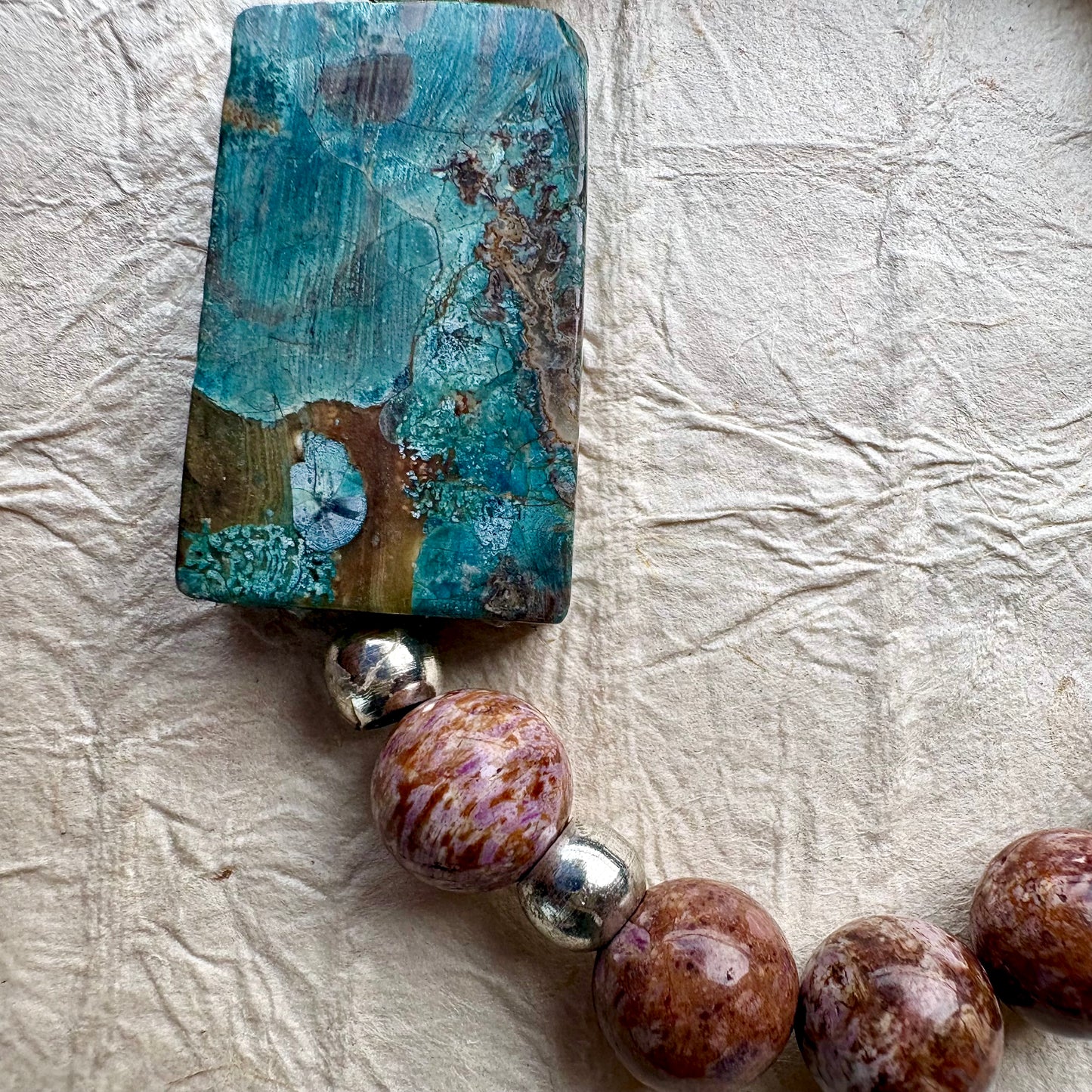 A close-up of a beaded jewelry piece featuring a large rectangular turquoise-blue stone pendant with brown and gold veining, connected to round brown and purple marbled beads separated by small silver spacer beads, laid on a textured off-white surface.