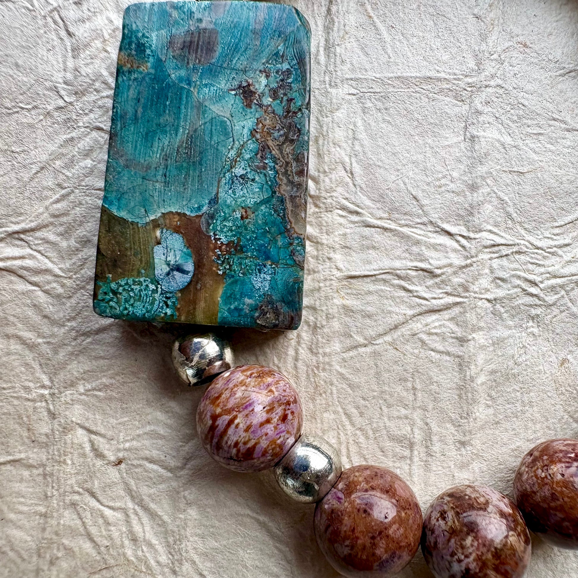 A close-up of a beaded jewelry piece featuring a large rectangular turquoise-blue stone pendant with brown and gold veining, connected to round brown and purple marbled beads separated by small silver spacer beads, laid on a textured off-white surface.