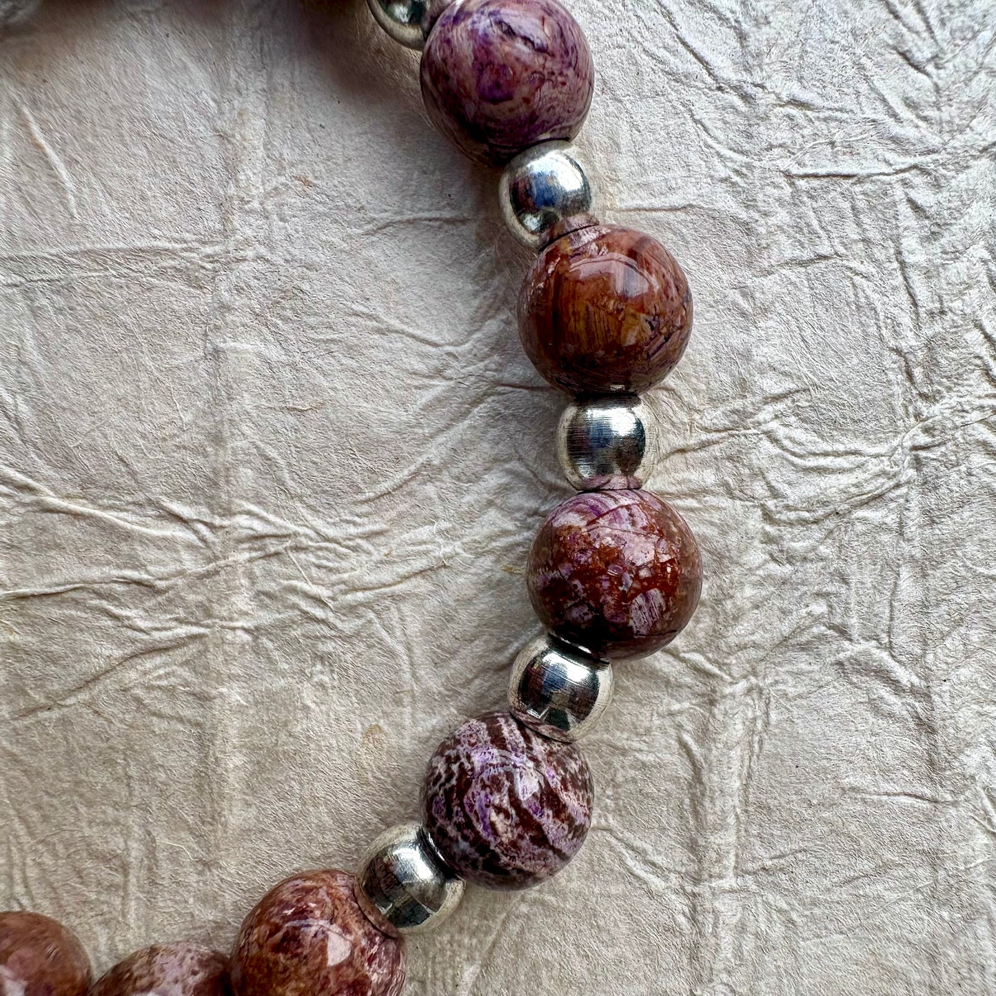 A close-up of a beaded strand featuring polished brown and purple marbled stone beads alternating with shiny silver metallic beads, laid on a textured beige surface.