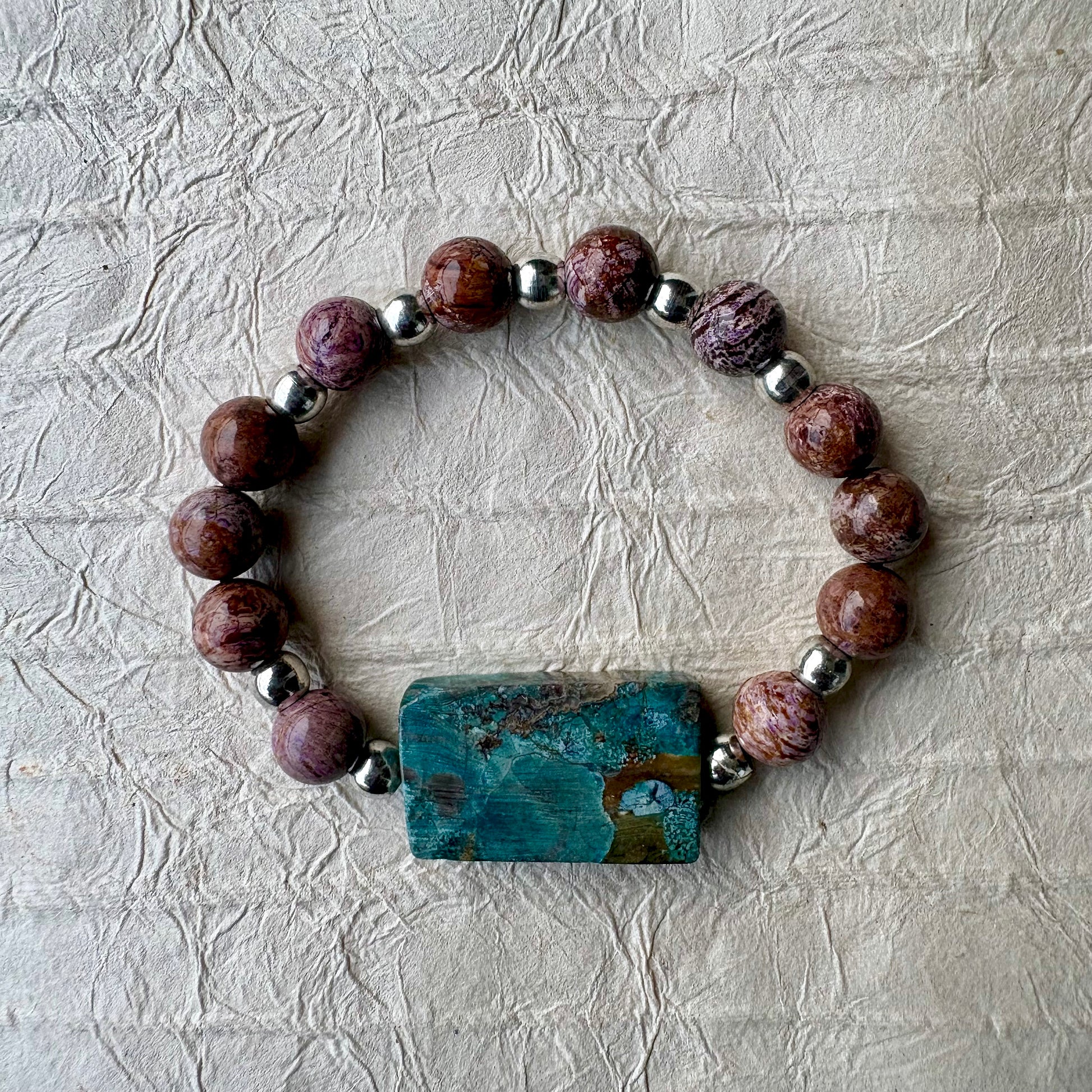 A beaded stretch bracelet featuring round brown and purple jasper beads, polished silver spacer beads, and a rectangular turquoise-blue stone centerpiece with natural matrix patterns, displayed on a textured off-white paper background.