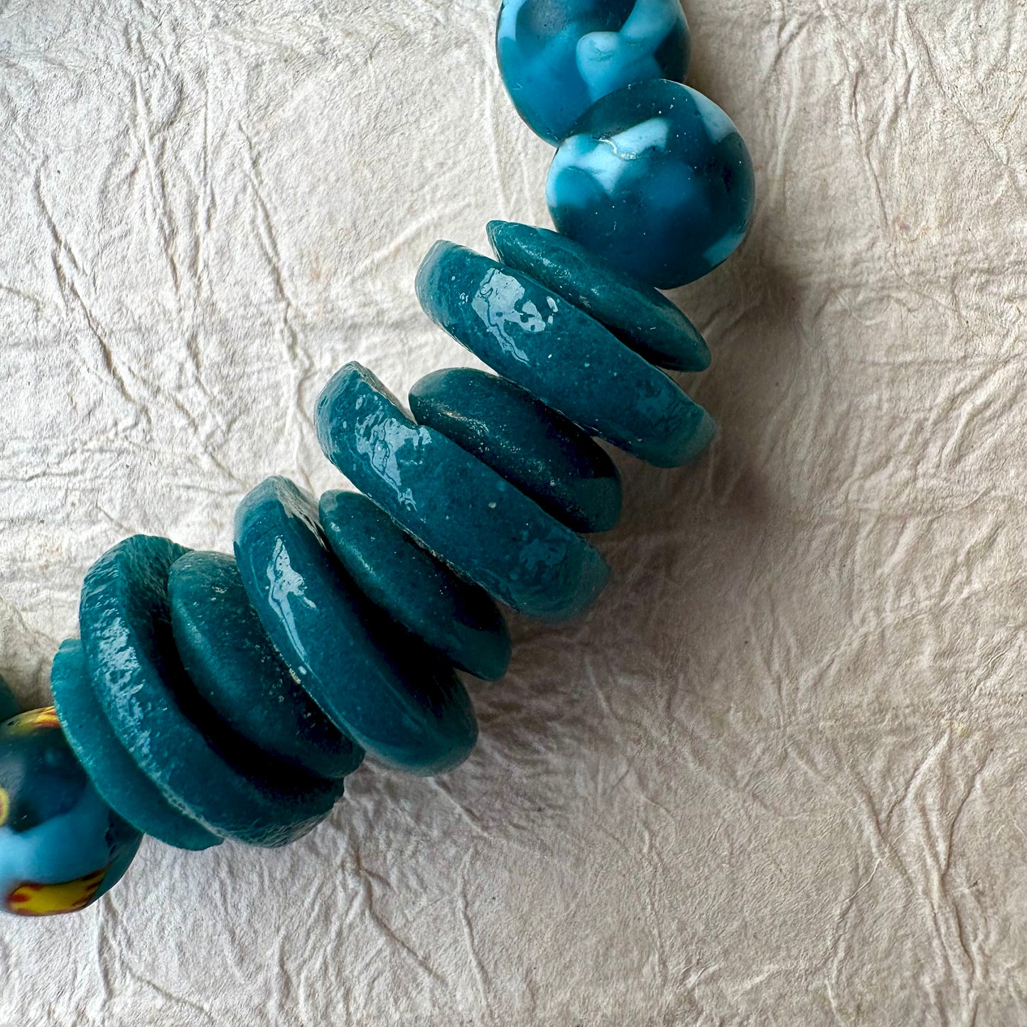 A close-up of a beaded strand featuring glossy, stacked teal-blue disc beads and a few larger marbled blue and white beads, laid diagonally on a textured off-white paper background.