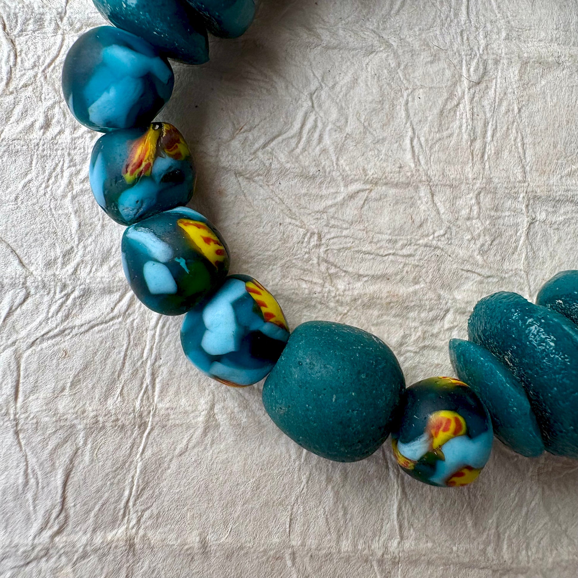 A close-up of a handmade beaded necklace featuring large, rounded marbled beads in teal, blue, yellow, and white, arranged on a textured off-white paper background.