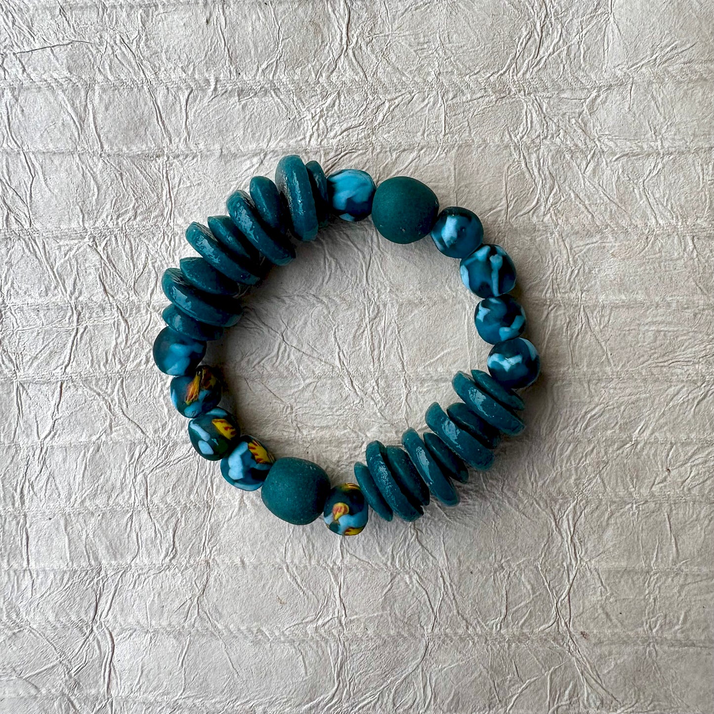 A handmade beaded stretch bracelet featuring a mix of matte teal-green beads, glossy marbled blue-green beads, and textured dark teal disc beads, displayed on a textured light gray paper background.
