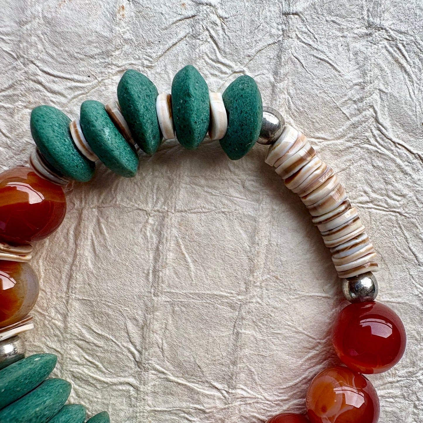 A close-up of a handmade beaded bracelet featuring textured green stone discs, polished red agate beads, and a section of cream-colored spiral shell beads, displayed on a textured light gray background.