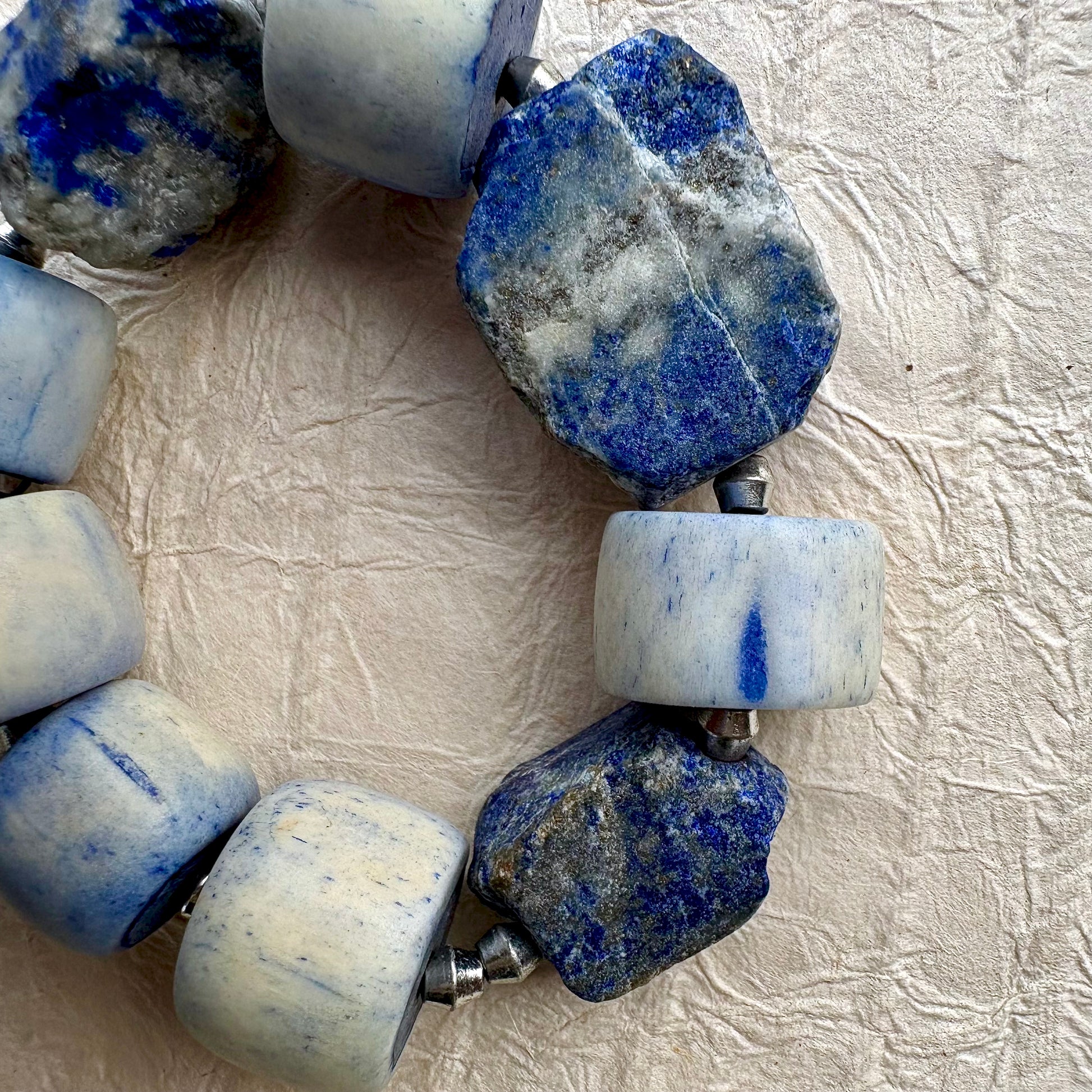A close-up of a beaded necklace featuring alternating smooth off-white beads with blue marbling and rough, polished blue stone beads, displayed on a textured beige fabric background.
