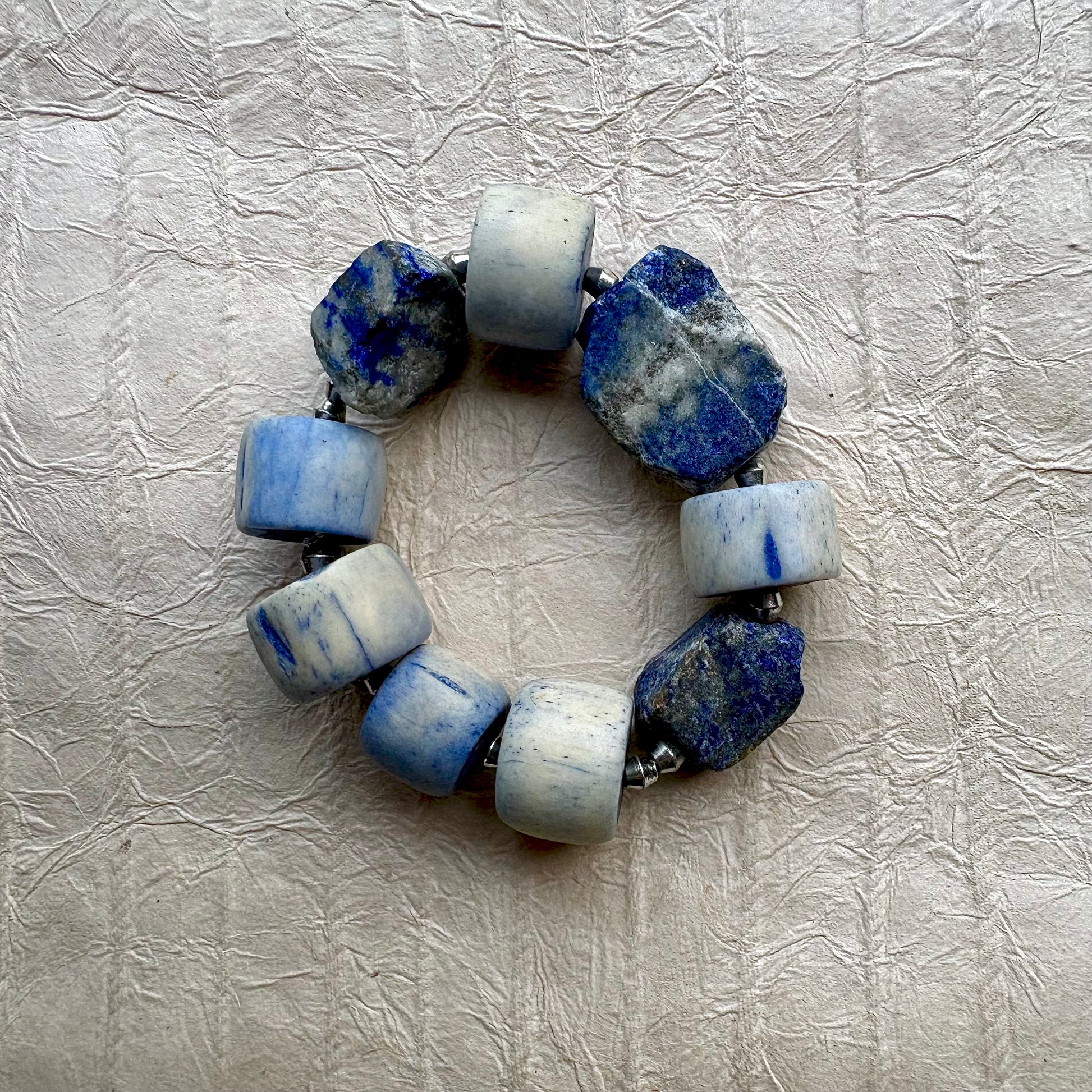A beaded stretch bracelet made of alternating smooth light blue and white cylindrical beads and rough blue stone slices, displayed on a textured beige fabric background.